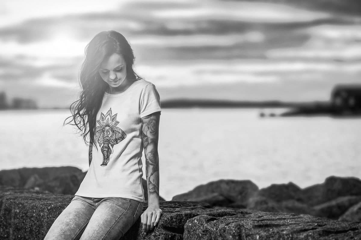 Kvinne i T-skjorte med elefanttrykk sitter på en stein ved havet, i svart-hvitt